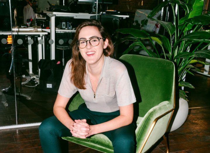 Katie Mannion sits on a green velvet chair in front of a leafy green plant. She is leaning forward with her elbows on her knees, smiling broadly.