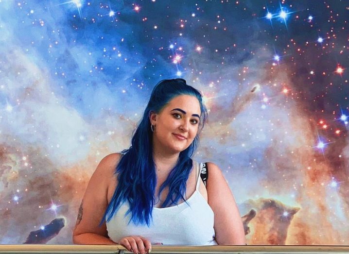 A young woman with dyed blue hair smiles at the camera in front of a backdrop showing a starry sky.