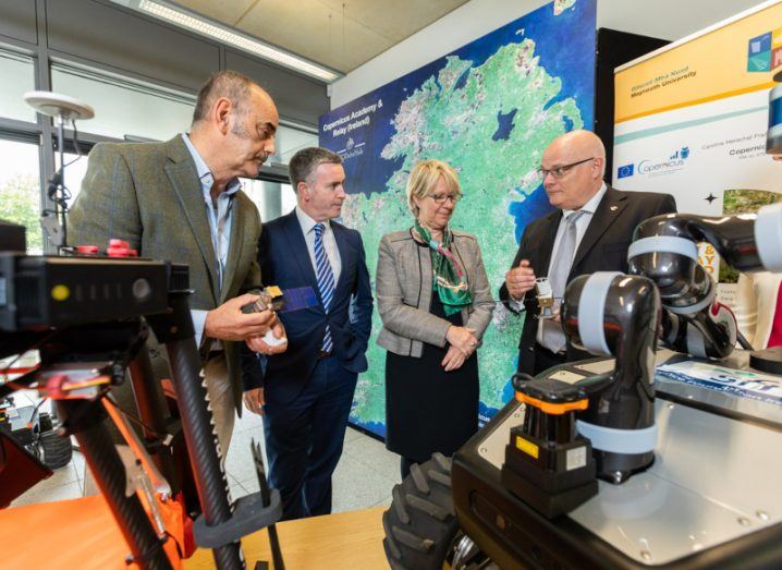 Photograph of three men and one woman looking at a device, with a map of Ireland in the background. They are Prof Tim McCarthy, Damien English, TD, Prof Eeva Leinonen and Conor Sheehan.