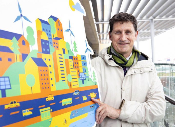 Eamon Ryan stands beside a large cardboard cutout of a cityscape illustration.