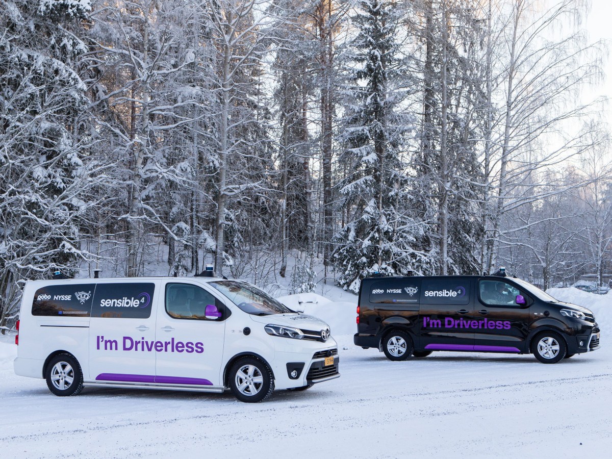 Two self-driving Toyota Proace vehicles, one white and one black, on snowy terrain with trees in the background. Both vehicles have 'I'm Driverless' written on the side in purple.