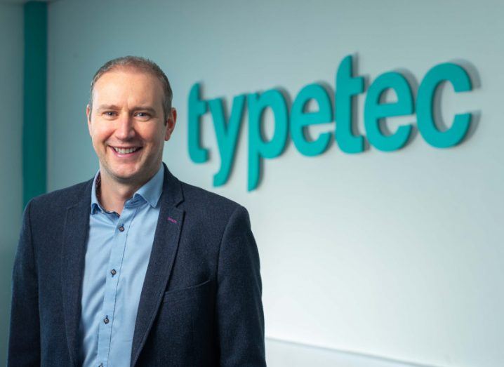 Paul Dooley stands in an office space in front of a wall that says Typetec on it.