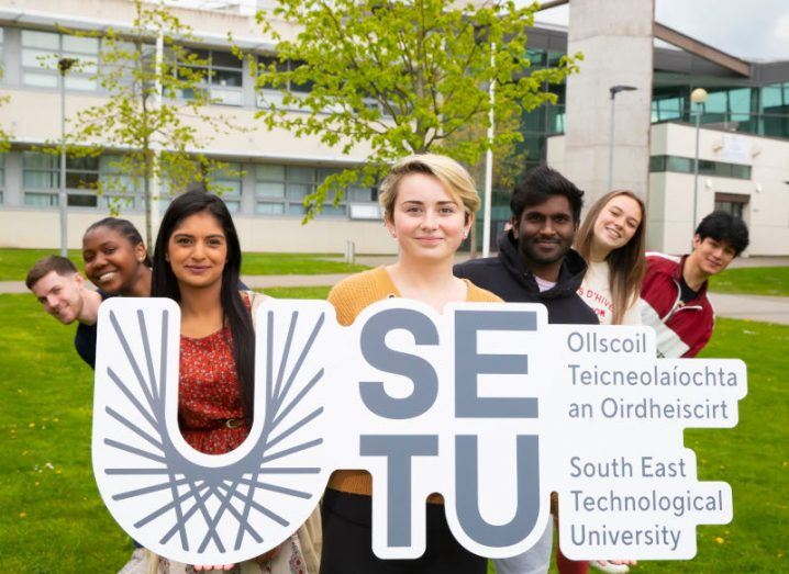 Seven students from SETU holding a sign with the university's name and logo outside standing in front of a building on a patch of grass.