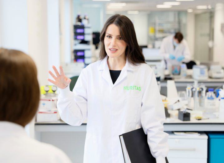Nuritas CEO Dr Nora Khaldi standing in a lab wearing a lab coat talking to somebody only partially visible.