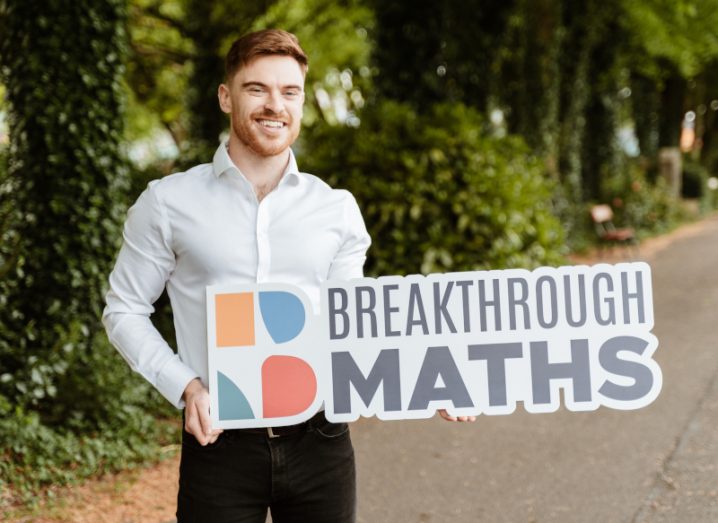 TJ Hegarty, founder of Breakthrough Maths, holding a sign with the company's branding on it. He is standing outside in a laneway with trees behind him.