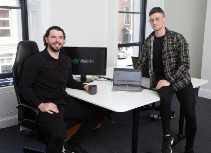 VisionR founders Oran Mulvey and Shane O'Sullivan sitting at a desk in a bright open office space. There is a computer on the desk between the two.