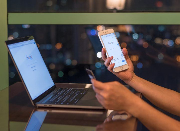 A person holding a smartphone with Google search open and a payment card in the other hand. A laptop is open in front with Google search displayed on screen.