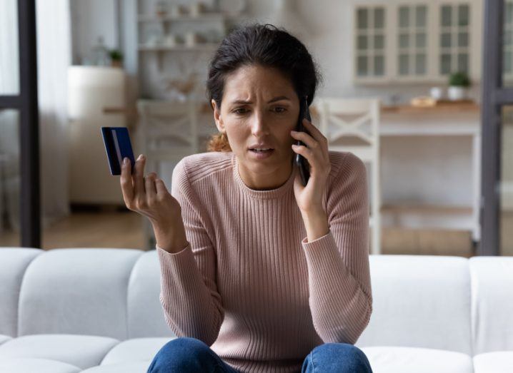 A woman is at home talking to someone on the phone while holding up her payment card.