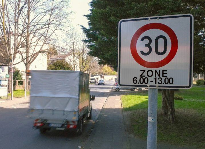 A speed limit sign with the number 30 on it. A mini truck can be seen driving past it on the road.