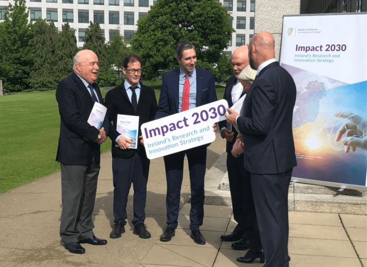 A group of people gather around Simon Harris, who is holding a sign that reads "Impact 2030".