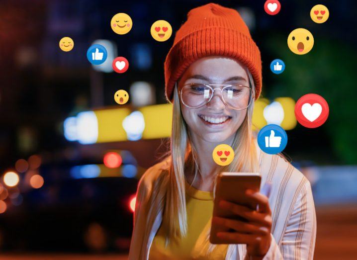 A woman wearing a beanie smiles at her smartphone as emoji reactions pop out of her phone.
