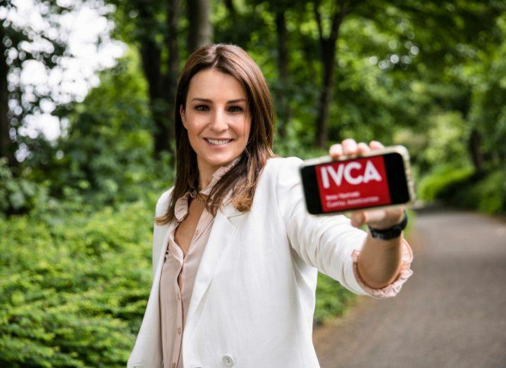 Nicola McClafferty stands in a park holding up a smartphone with the IVCA logo on a red background.
