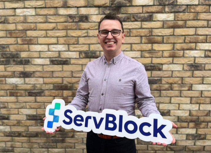 ServBlock CEO John Ward stands in front of a brick wall holding a ServBlock sign.