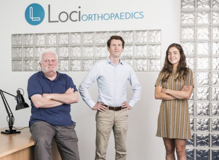 Photograph of two men and one woman smiling at the camera. One of the men is sitting on a table while the other two people are standing. The Loci Orthopaedics logo is behind them. They are Gerry Clarke, Dr Brendan Boland and Fiona Mangan.