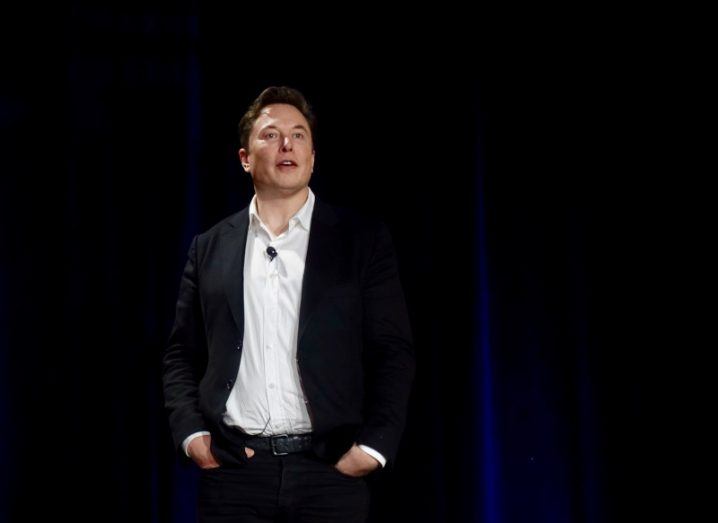 Elon Musk in a black suit jacket and a white shirt with his hands in his pockets, speaking on a dark stage.