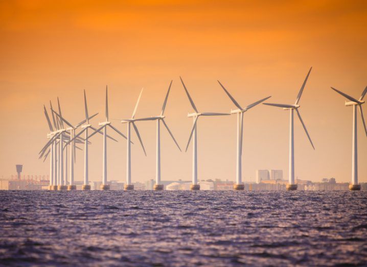A group of offshore wind turbines in the sea, with an orange sky and a city in the background.