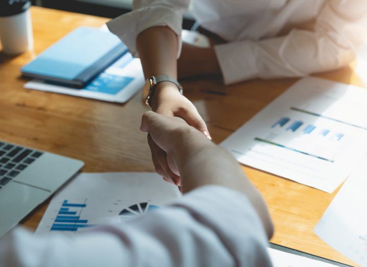 Business people shaking hands across a table with graphs and a laptop in the background.