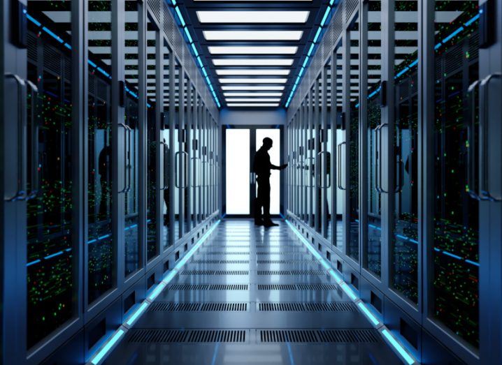 Man in the end of a data centre hallway, with cloud servers on either side of the image. The man is standing down the hallway with a white light coming from a door behind him.