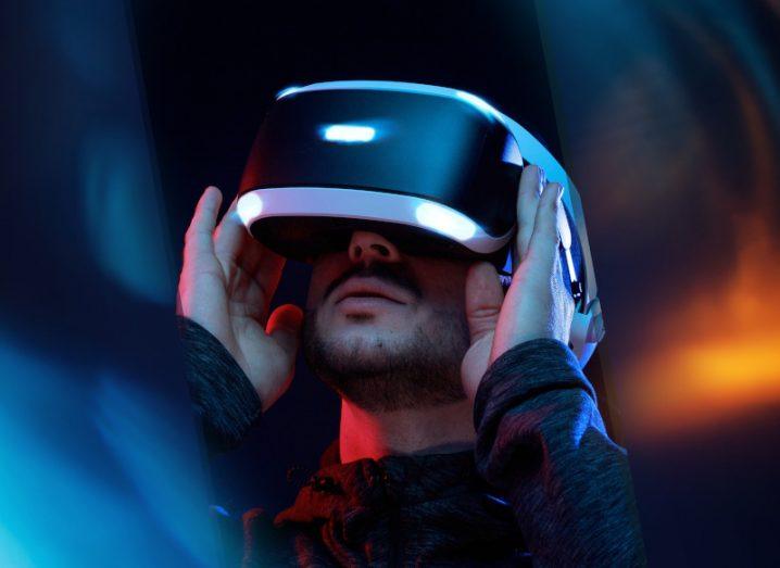 Man wearing a virtual reality headset and holding the sides with his hands, in a dark background with blue lighting to the left and orange lighting to the right.