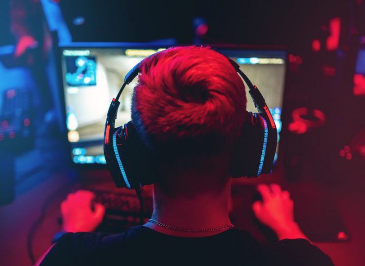 The back of a person's head, wearing a headset and looking at a PC monitor while gaming, with red and blue lighting.