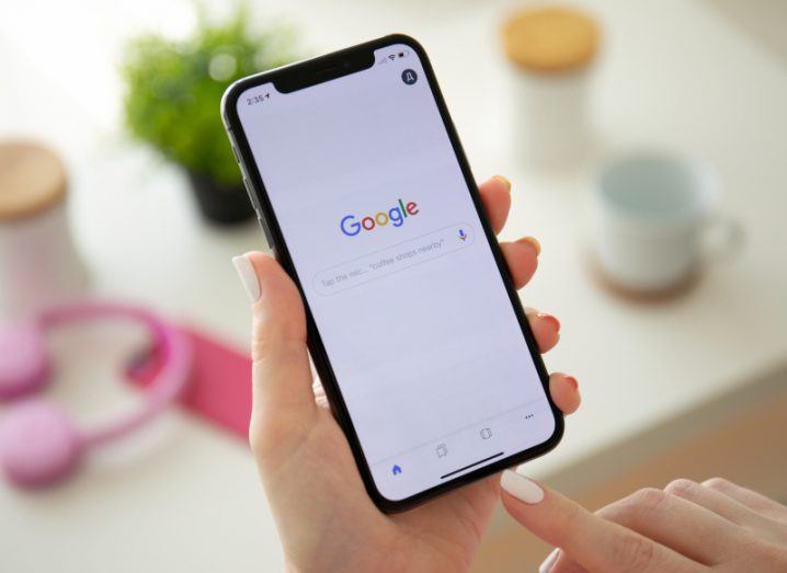 Person holding an iPhone which has a Google search engine on the screen. There is a table in the background with a plant and a cup resting on it.