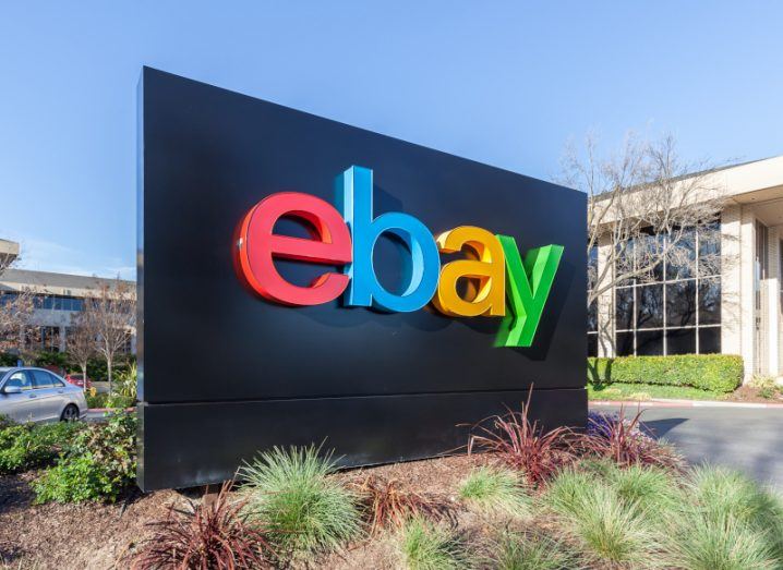 eBay logo on a black board in front of a building, with a road behind the sign and small green plants in front. A clear blue sky is visible.