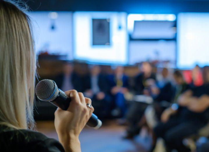 Business woman addressing crowd of people in a room using microphone.