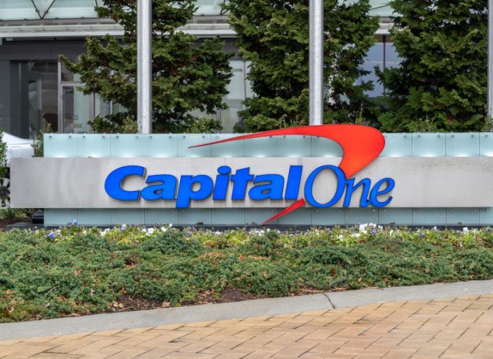 Capital One logo in front of a building, with trees behind the logo and grass in front of it. There is a brown and grey path in the front of the image.