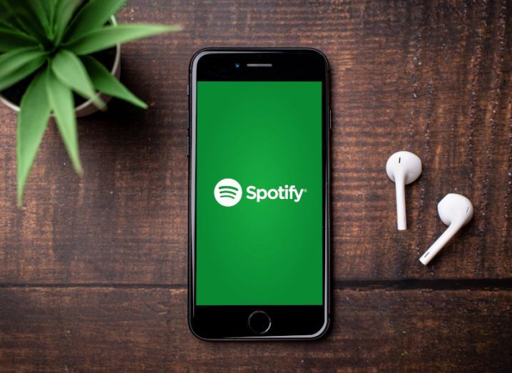 Spotify logo written in white on a green background on a mobile phone screen. The phone is laying on a dark brown wooden table, with a pair of wireless earphones and a small houseplant next to it.