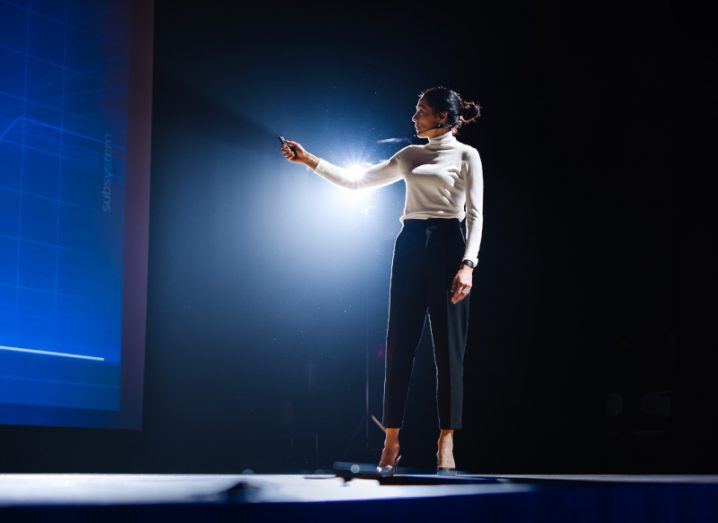 A businesswoman in a leadership role presenting something on stage in semi-darkness.
