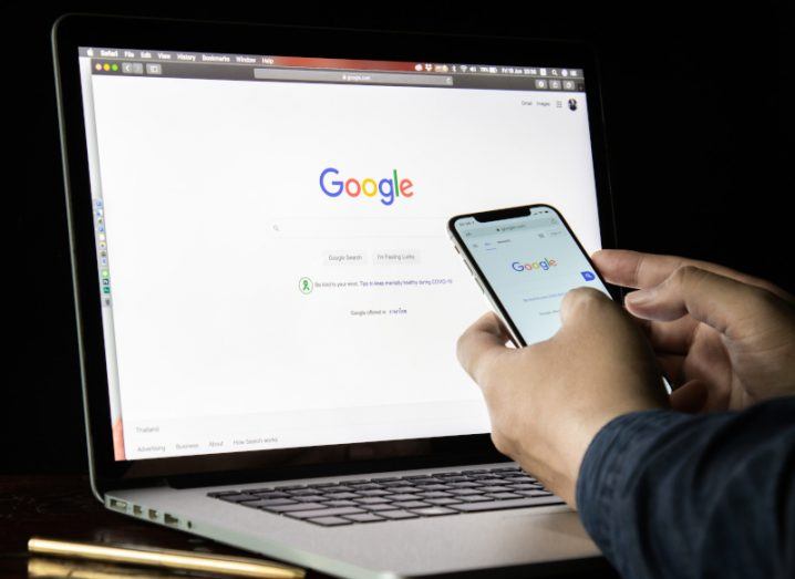 A person's hands holding a mobile phone with the Google logo and a search bar. A similar search icon is on a laptop screen behind the phone.