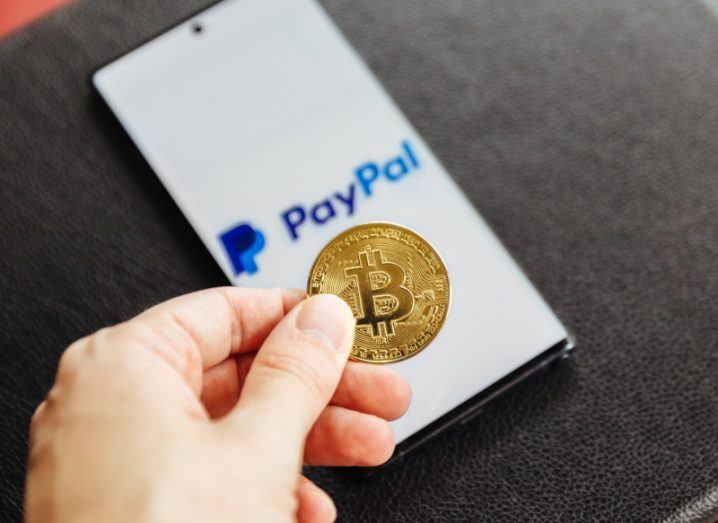 A hand holding a gold coin with the bitcoin logo on it. A smartphone is laying on a table behind the coin displaying the PayPal logo on a white background.