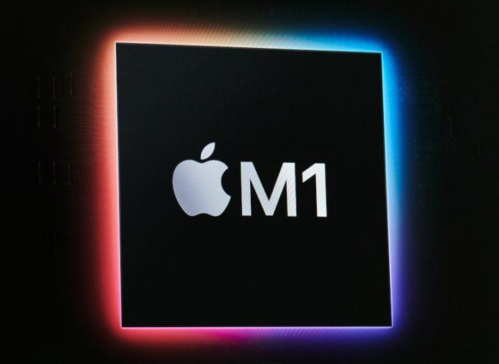 Apple logo next to M1, which is a type of processor chip, on a black square in a dark background. The square has a multi-coloured outline, with blue on the right and red on the left.