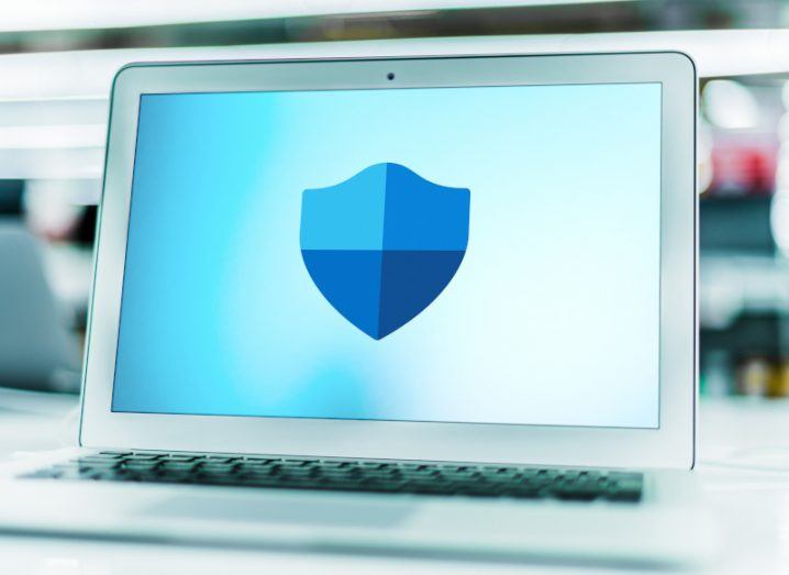 Laptop screen displaying the blue Microsoft Defender logo, on a blue and white background. The laptop is resting on a white table.