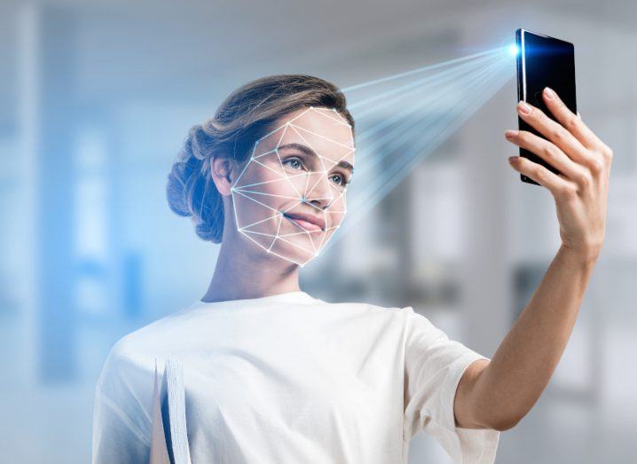 Woman wearing a white blouse holding a mobile phone with a beam of light scanning her face, representing facial recognition technology.