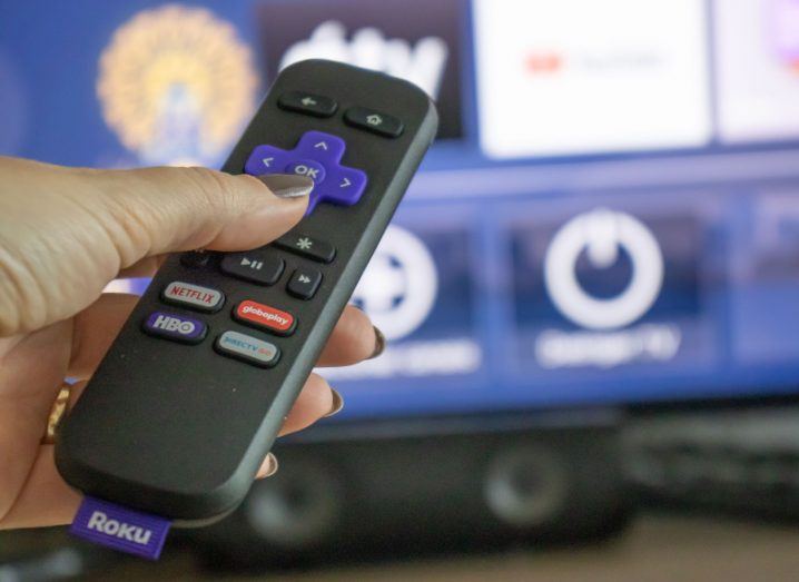 Roku TV streaming remote control being held in a person's hand. A TV is visible in the background.