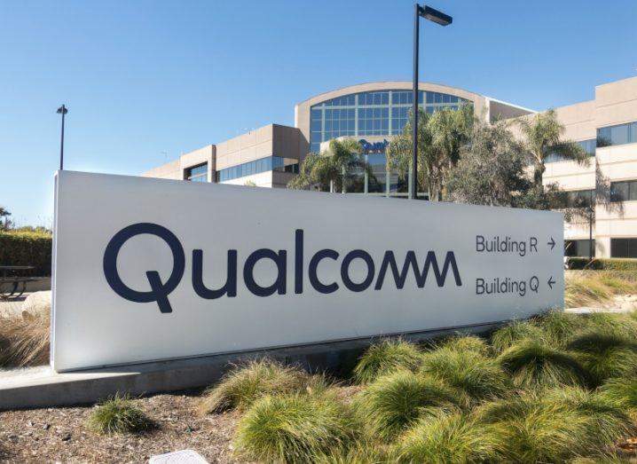Qualcomm logo on a white billboard with some green plants in front of it and a building in the background. Building R and Building Q are written on next to the logo, while a clear blue sky is visible.