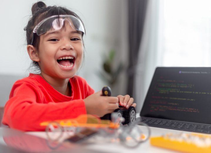 Child sitting in front of computer with coding language on its screen wearing lab goggles.