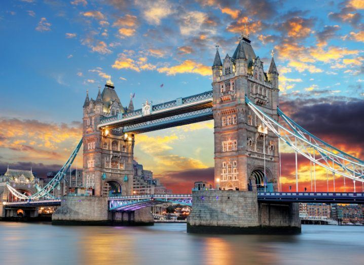 The Tower Bridge in London, with calm blue water under it and a blue sky above it. There are orange clouds in the sky as a result of a sunset.