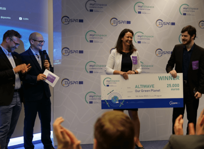 Two people stand holding a large cheque in a room of people clapping.