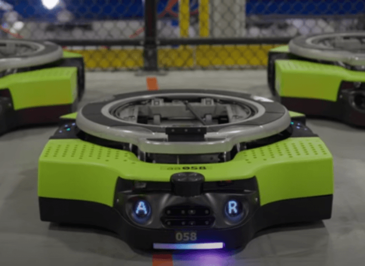 Three green robots on a warehouse floor. The front robot has a blue light at the bottom, with the letters A and R lit up on its front. The robot is called Proteus, which Amazon said is its first fully autonomous mobile robot.