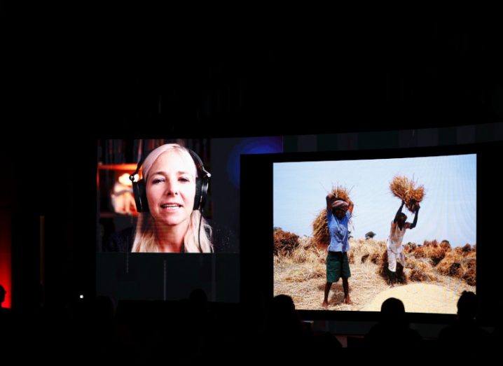 A woman on a screen wearing headphones speaking to an audience, with a photo on the screen of two farmers. She is Prof Alice Roberts speaking at Future Human 2022.