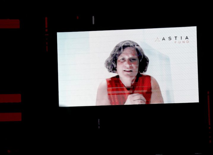 Sharon Vosmek appearing in head and shoulders view on a large screen in a dark room with the Astia logo on her screen.