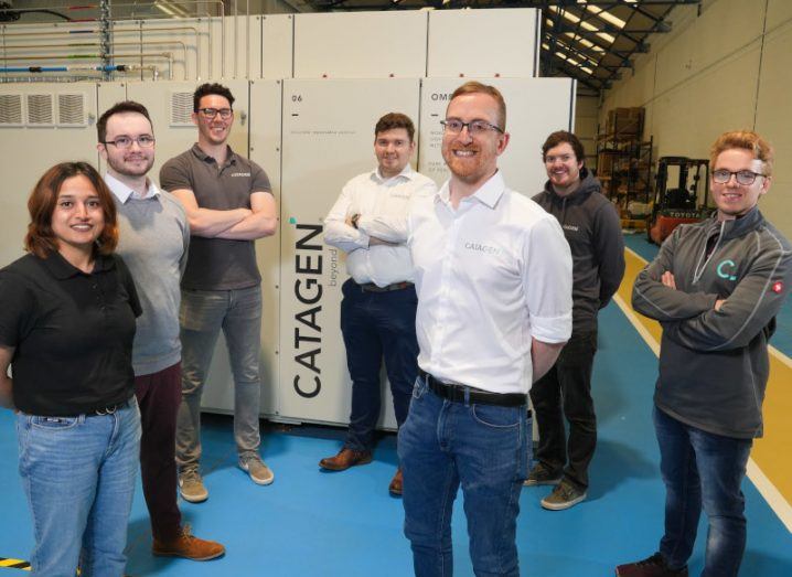 Seven people standing and smiling in a warehouse, with the Catagen logo behind them on the side of a large appliance.