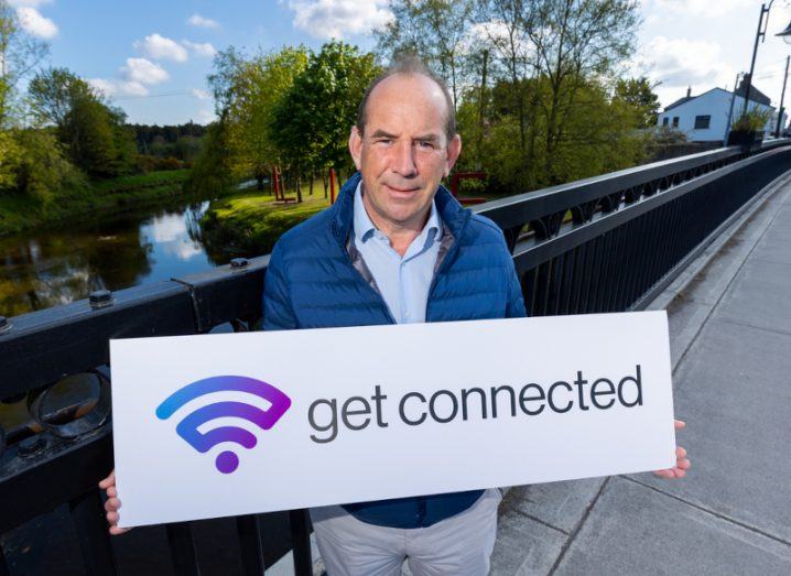 A man holding a sign that reads "get connected" with a wi-fi symbol on it. The man is standing on a bridge that is over a river, with trees and a blue sky in the background. He is Colin Cunningham, managing director of Cellnex Ireland.