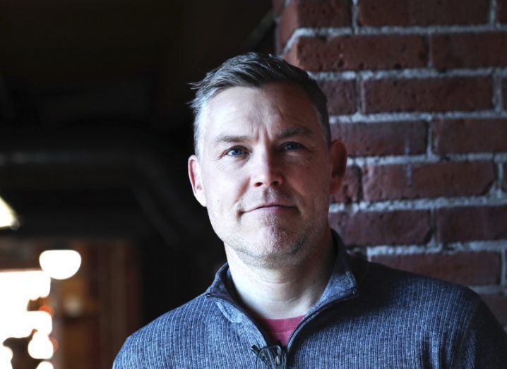 Brian Kastelein dressed in a knitted zip-up pictured against an exposed brick wall.