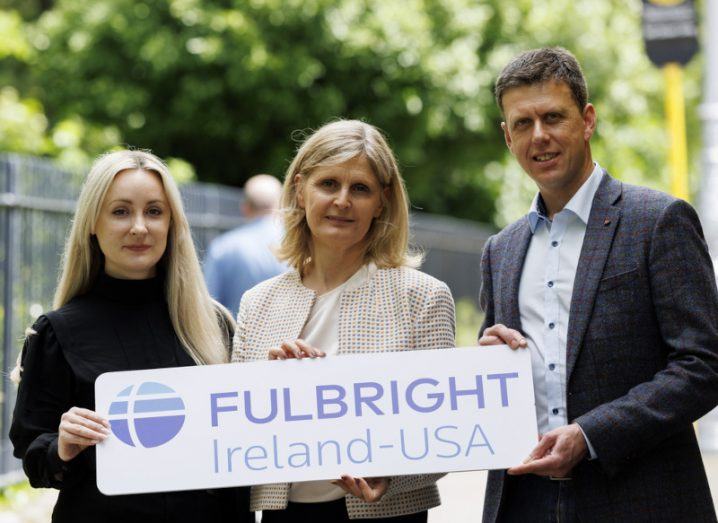 Two women and a man standing outside with green trees in the background. They are holding a sign that reads: Fulbright Ireland-USA.
