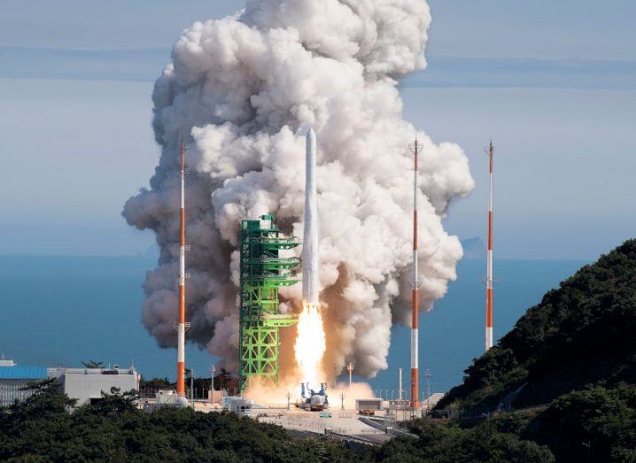 A rocket taking off from a launch site with a flame jet under it and a large cloud of smoke. The sea and the sky are visible in the distance.