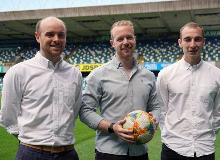 Photograph of three men smiling while standing in a sports pitch, with grass and seats visible behind them. The man in the center is holding a football in his hands. They are the founders of Pitchbooking.