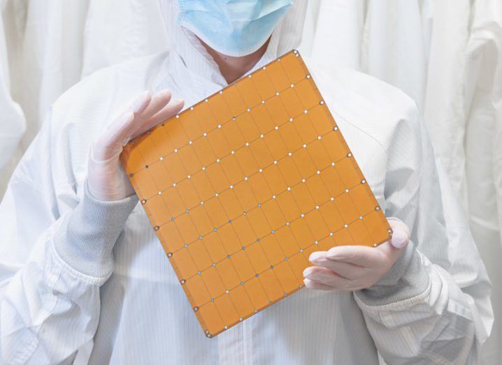 A person in a white lab outfit with a face mask and plastic gloves, holding a large brown processor. It is WSE-2 processor by Cerebras.